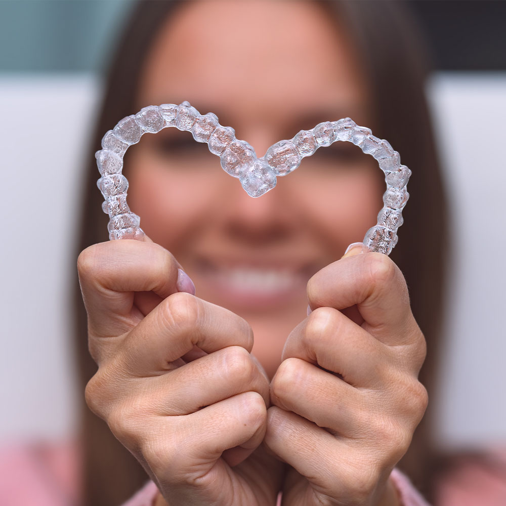 A woman is holding up a heart-shaped dental retainer with her hands, smiling at the camera.