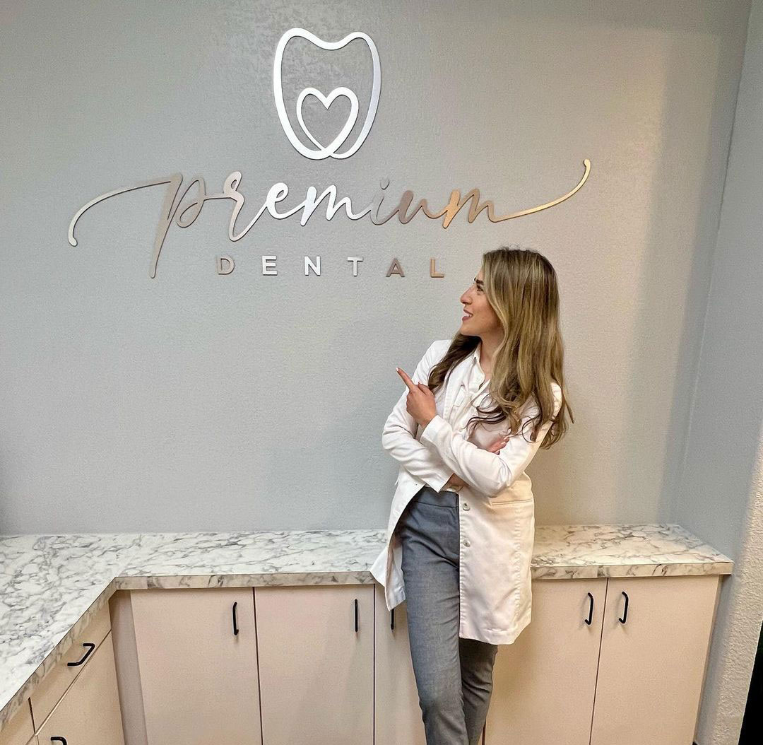 The image shows a woman standing in front of a sign for  Premium Dental,  which is likely the name of a dental practice. She appears to be posing for the photograph, with one hand on her hip and the other pointing towards the camera.