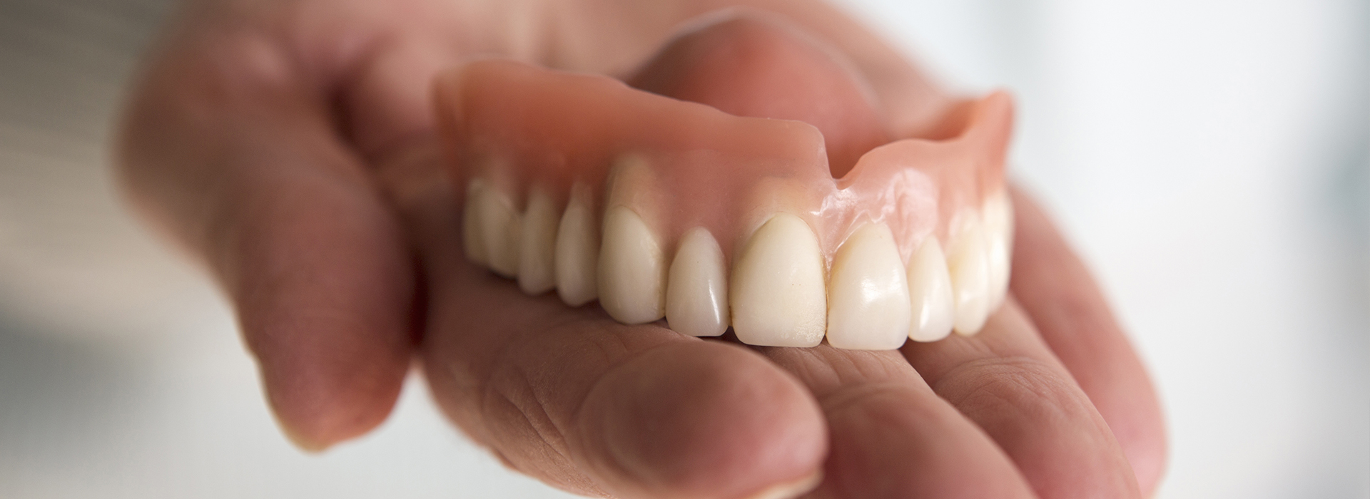 The image shows a person holding up a set of dentures, with the dentures prominently displayed against a neutral background.