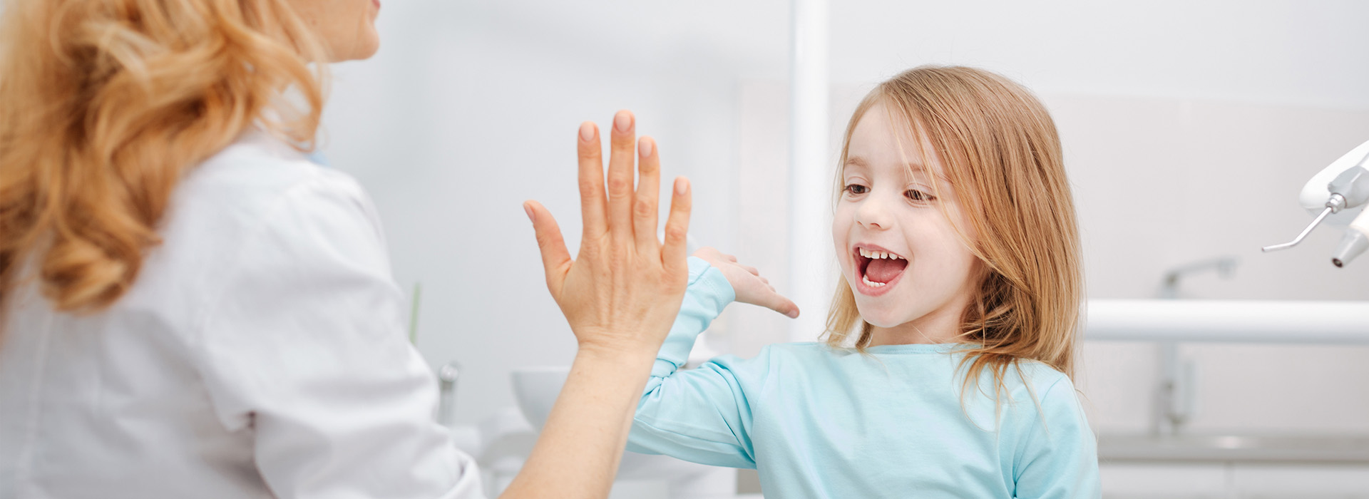 The image shows a young girl with long hair smiling at the camera while standing in front of a woman who is seated and appears to be holding her hand up, possibly in greeting or acknowledgement.